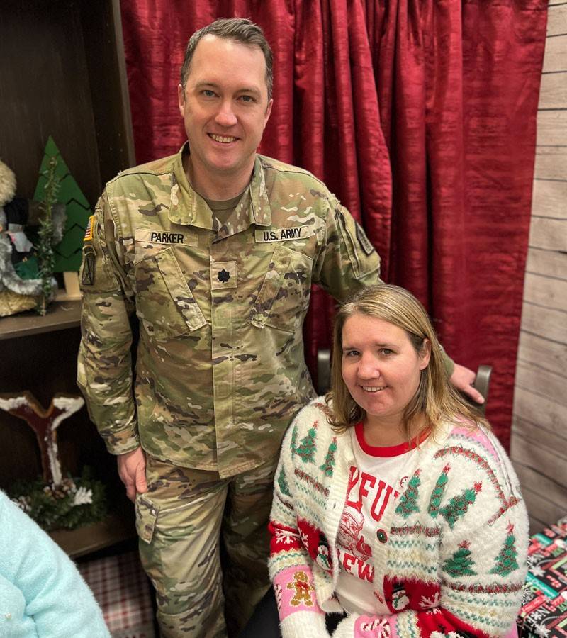 A Red Cross volunteer in a festive holiday sweater sits in front of a service member dressed in camo uniform.