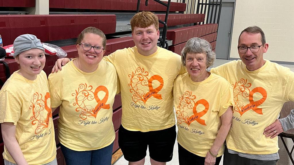 Five people in yellow shirts that read "Fight like Kaitlyn" smile at the camera.