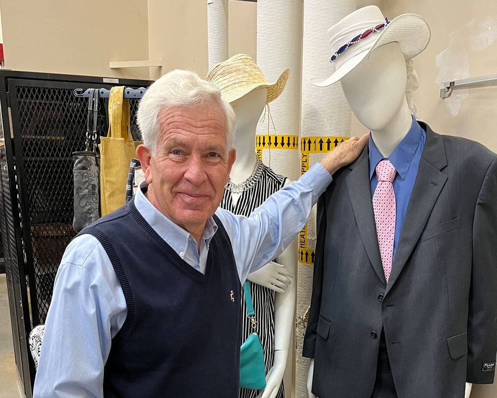 An American Red Cross volunteer smiles at the camera. They're dressed in a blue vest and shirt, with a Red Cross pin. They have their hand resting on a well-dressed mannequin.