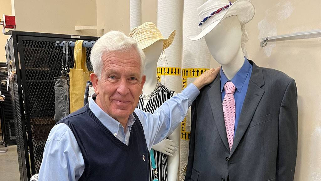 A person in a blue shirt and vest, wearing a Red Cross pin, stands with his hand on a well-dressed mannequin.