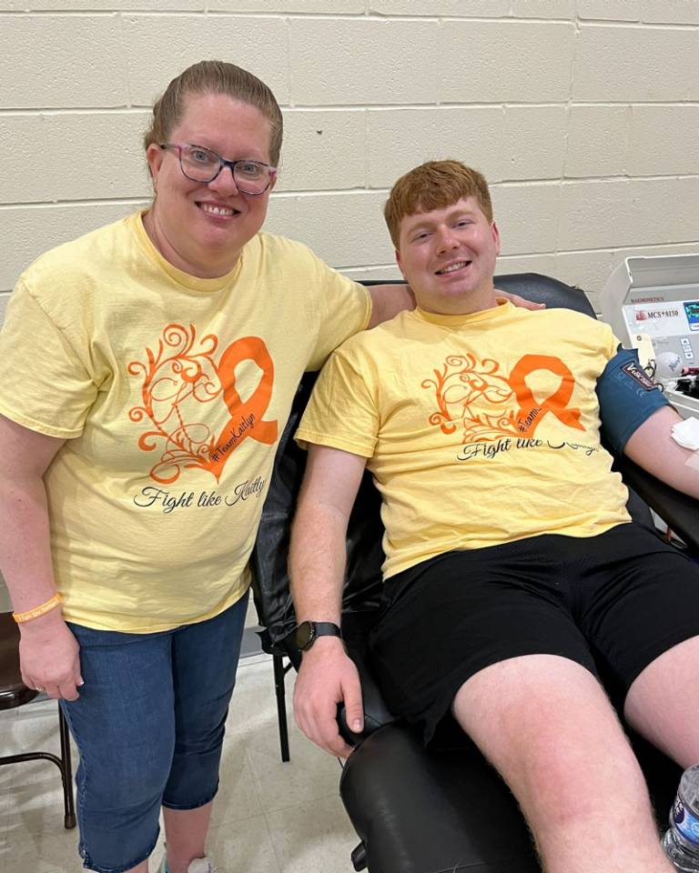 Two people in yellow shirts that read "Fight like Kaitlyn" smile at the camera. The person on the right is donating blood on a donation bed at a blood drive.
