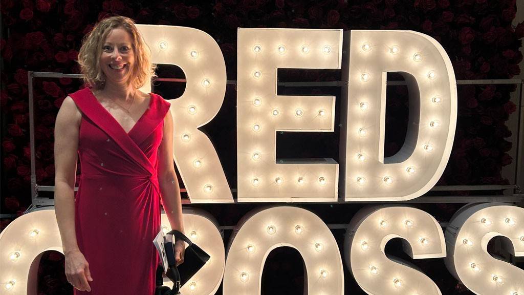  A person in a red dress poses for a photo in front of a large, lighted sign that reads "Red Cross".