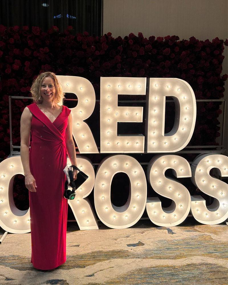 A person in a red formal dress stands in front of a lighted sign that reads "Red Cross". They are smiling at the camera.