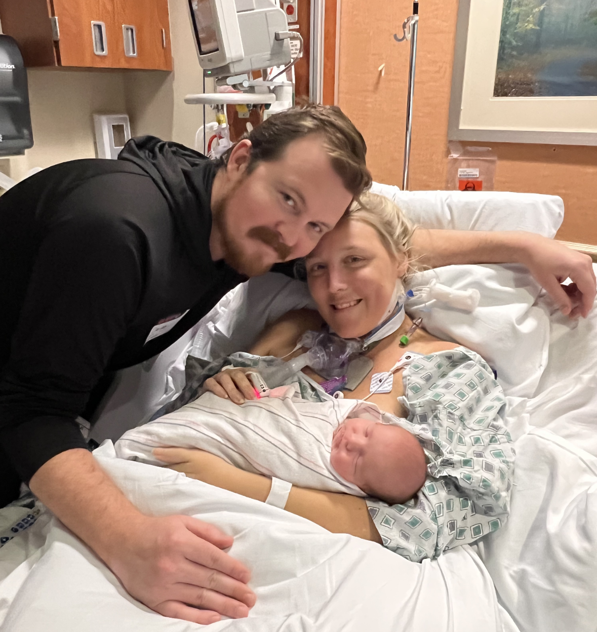 A person in a hospital bed with medical equipment and tubing attached to their chest holds a newborn. Another person in a black jacket stands next to them.