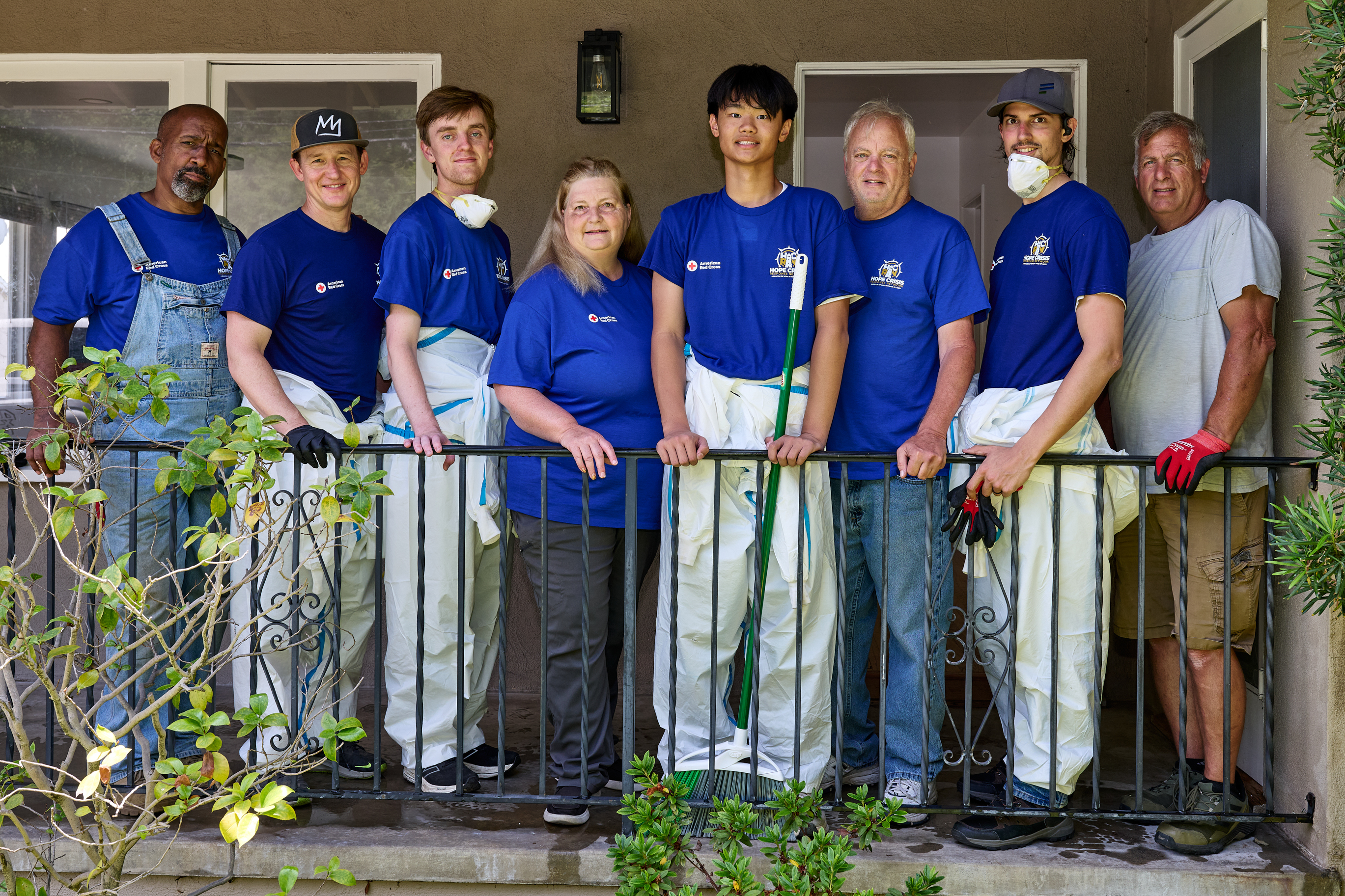 Group of 8 people wearing blue tee shirts and white protective jump suits standing in front of house railing