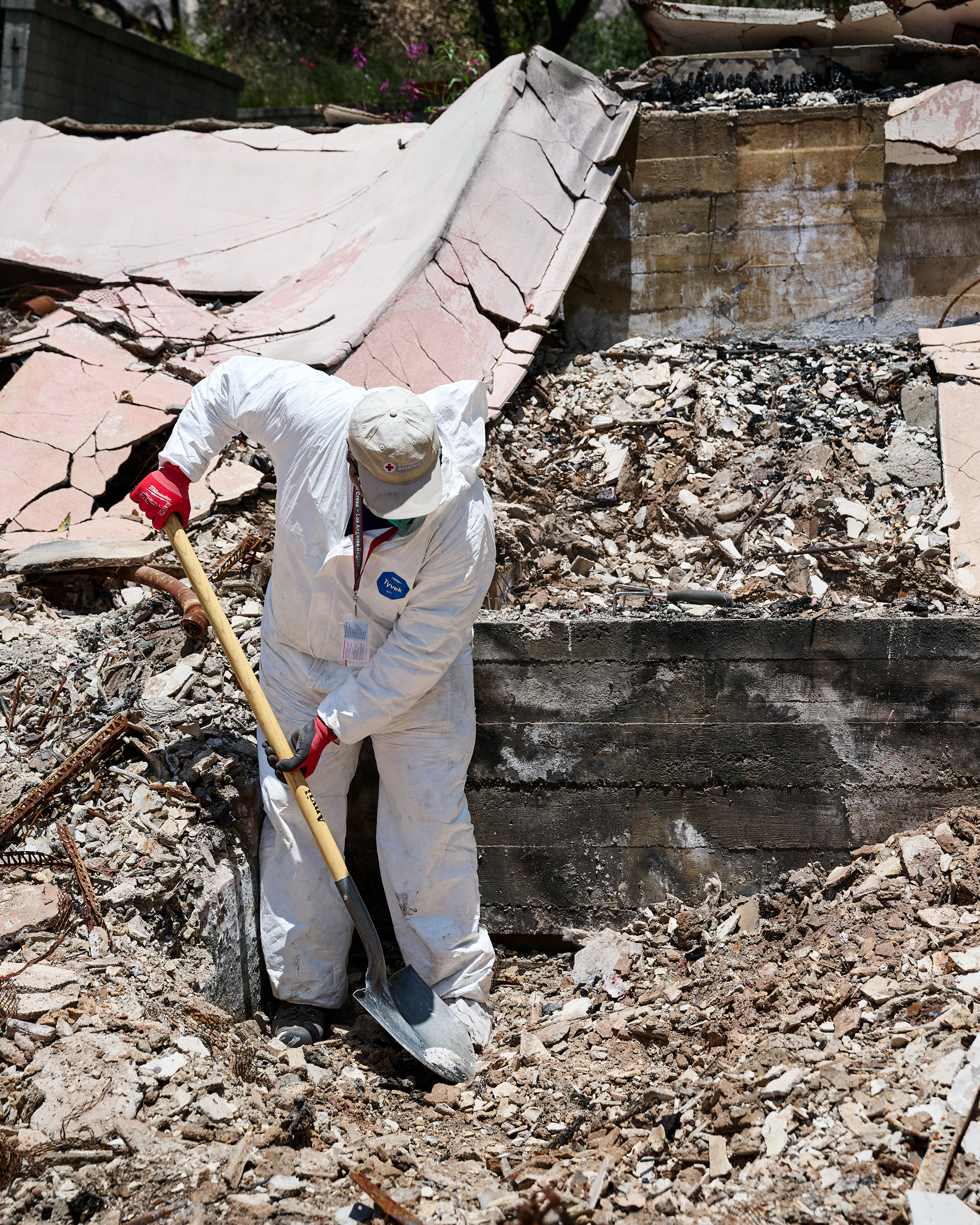 A man in a white protective jumpsuite and Red Cross logo baseball hat shovels fire debris