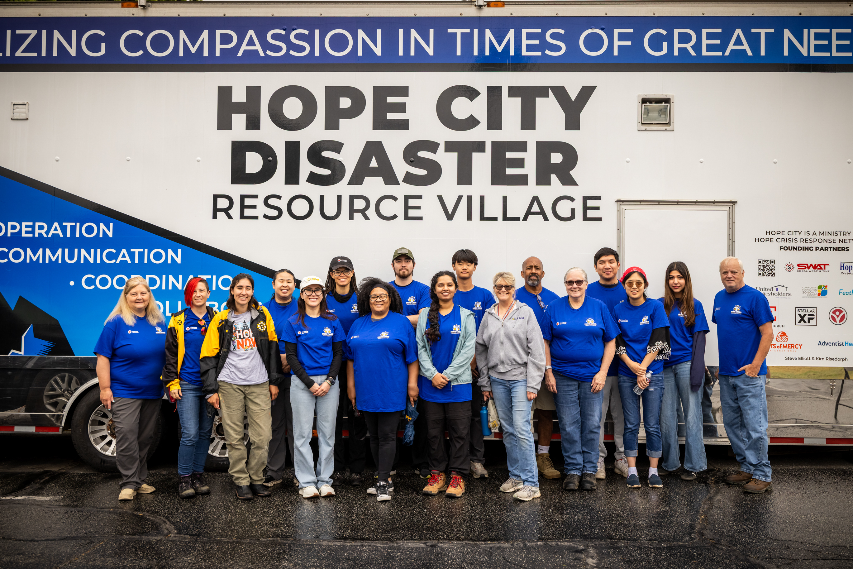 Group photo of 17 people wearing mostly blue tee shirts stand in front of a large truck that reads "Hope City Disaster Resource Village" on the side