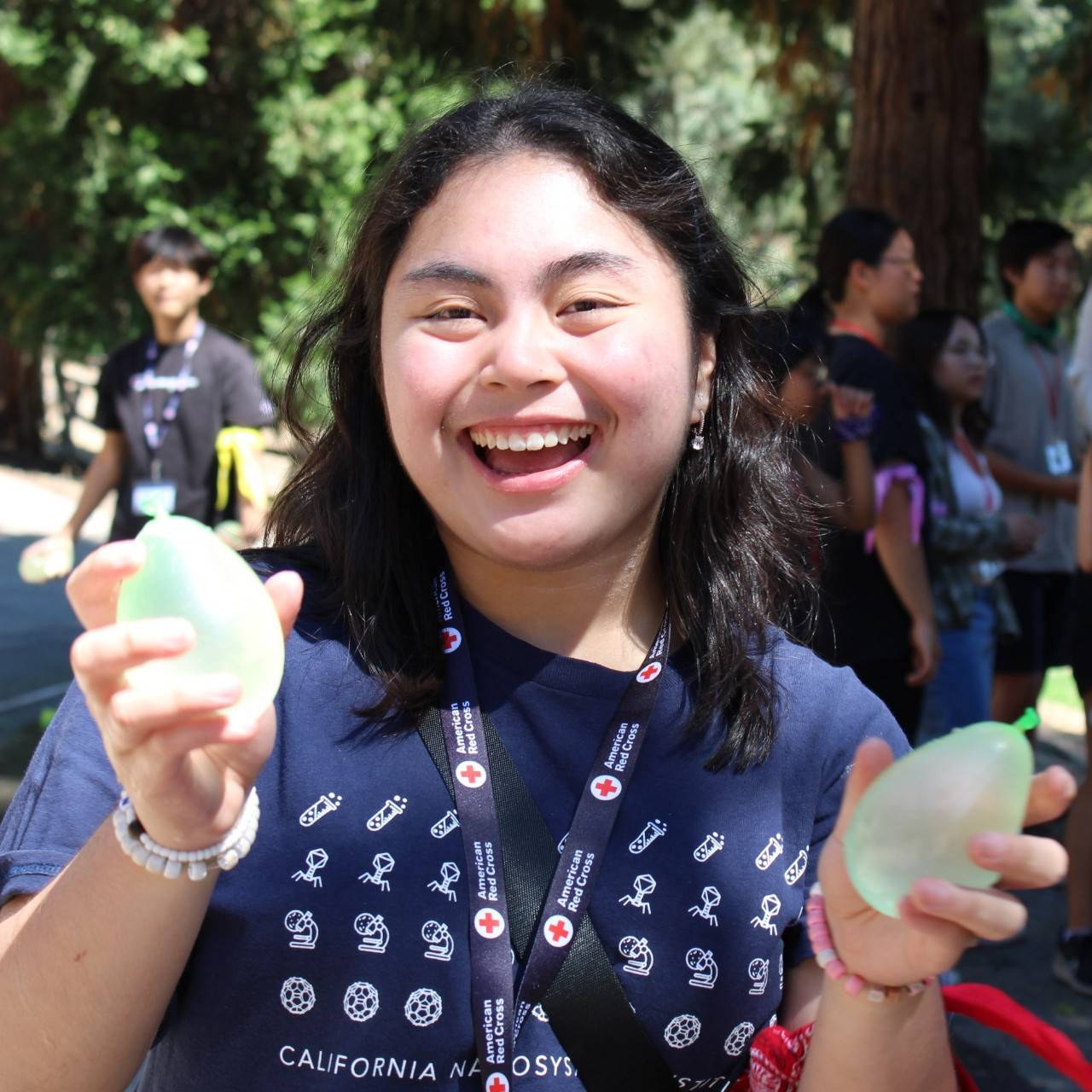 Smiling teenage adult with black hair holding one green water balloon in each hand