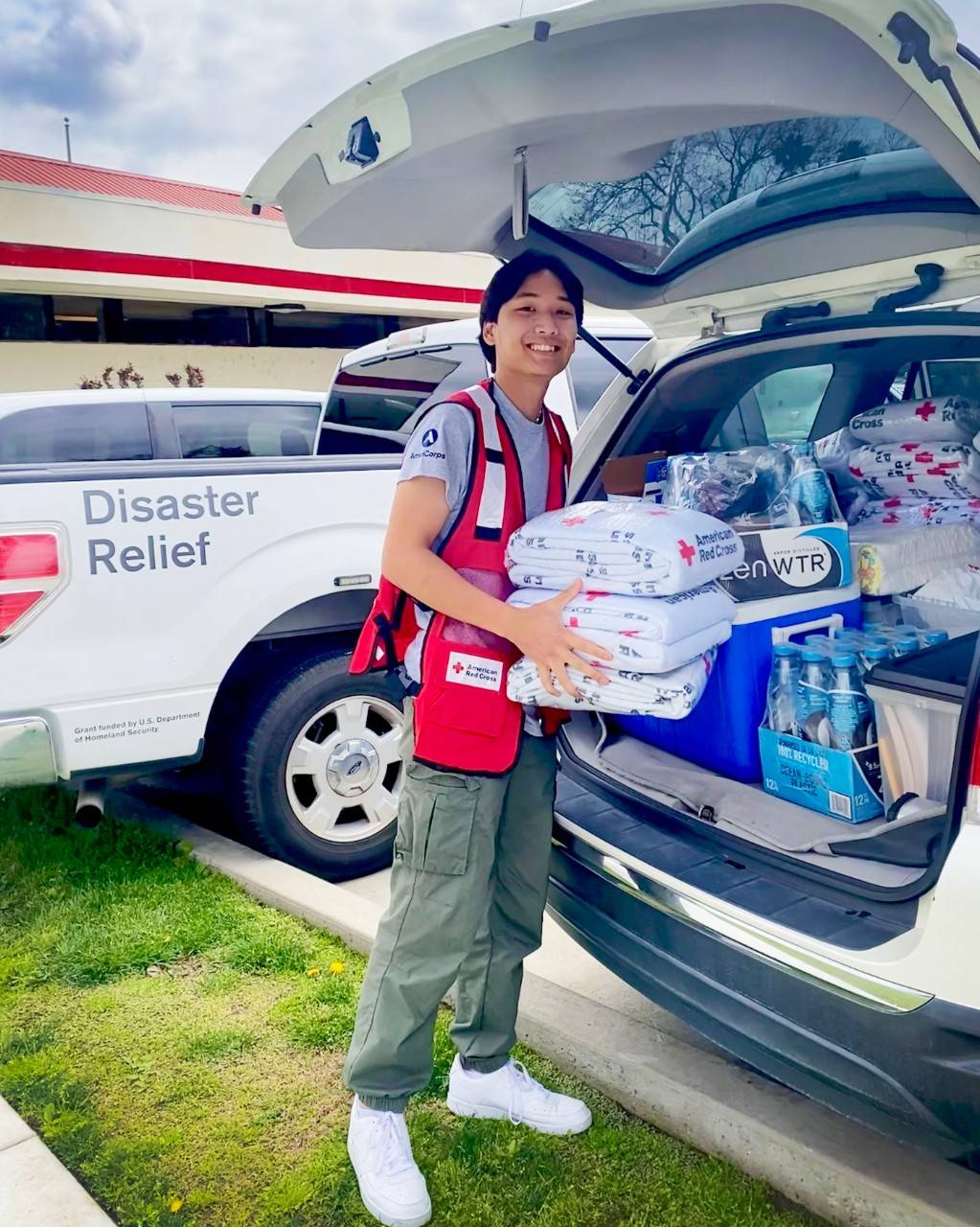 Red Cross volunteer wearing a red cross vest holding wrapped red cross blankets at the back of a red cross vehicle filled with supplies.