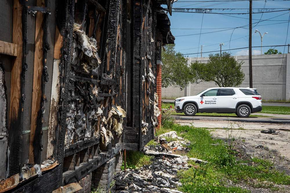 A Red Cross vehicle parked on the street next to a fire damaged house.