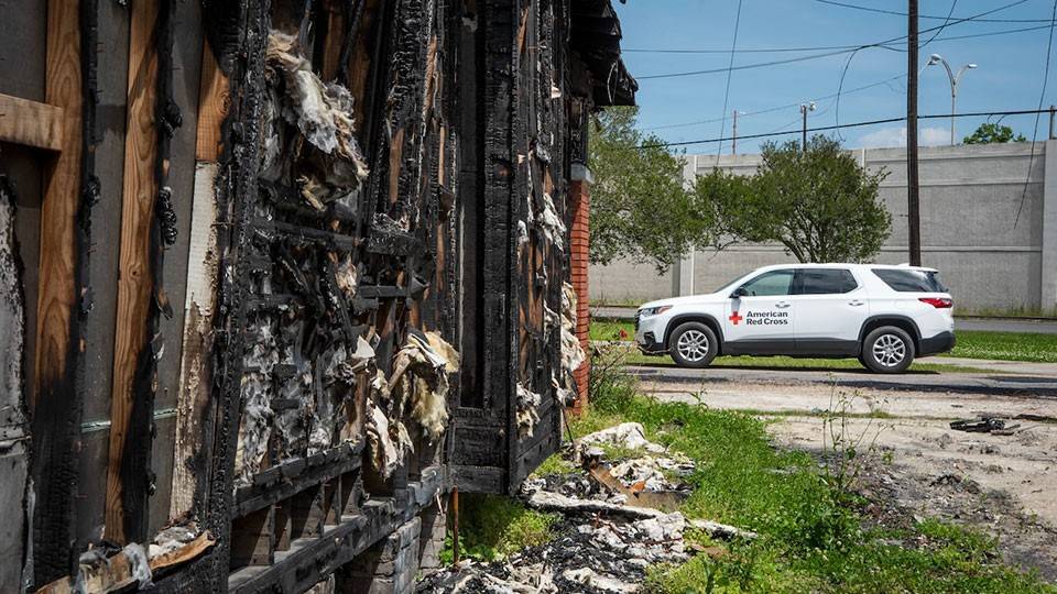 A Red Cross vehicle parked on the street next to a fire damaged house.