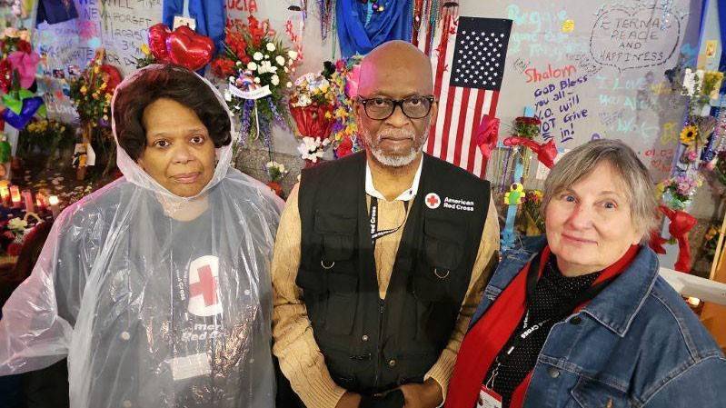 Dr. LeBan and two other Red Cross volunteers standing next to a memorial table with candles, flowers, balloons and signs.