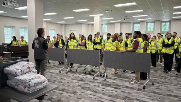 A group of people in bright yellow vests attentively listens to a speaker in a spacious, well-lit room.