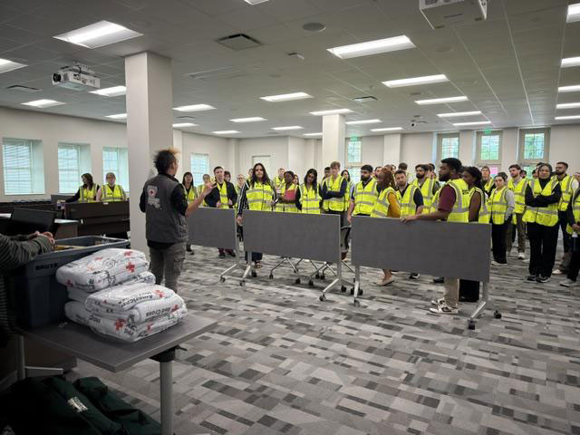A group of people in bright yellow vests attentively listens to a speaker in a spacious, well-lit room.