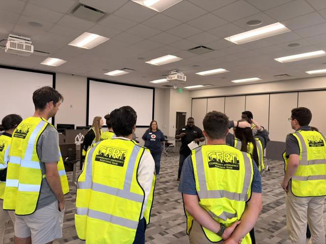 A group of people wearing yellow CERT vests stand in a classroom, attentively listening to instructors at the front.