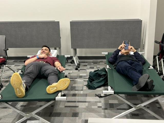 Two people relax on green cots in a room with gray walls and folding tables. One smiles, while the other uses a phone.