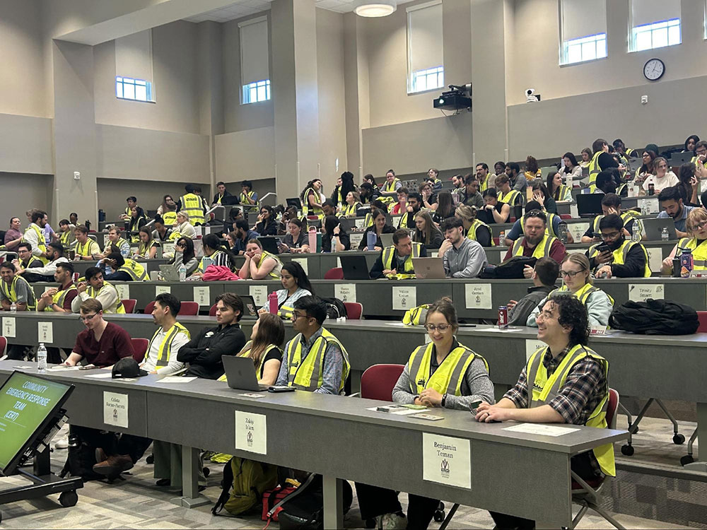 Lecture hall filled with attentive students wearing yellow safety vests. Many use laptops.