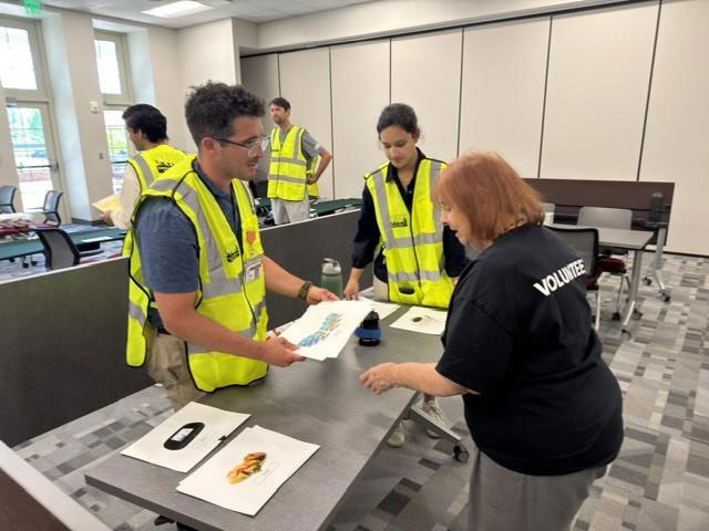 Two people wearing yellow safety vests and discussing papers with a volunteer in a black shirt in a classroom-like setting.
