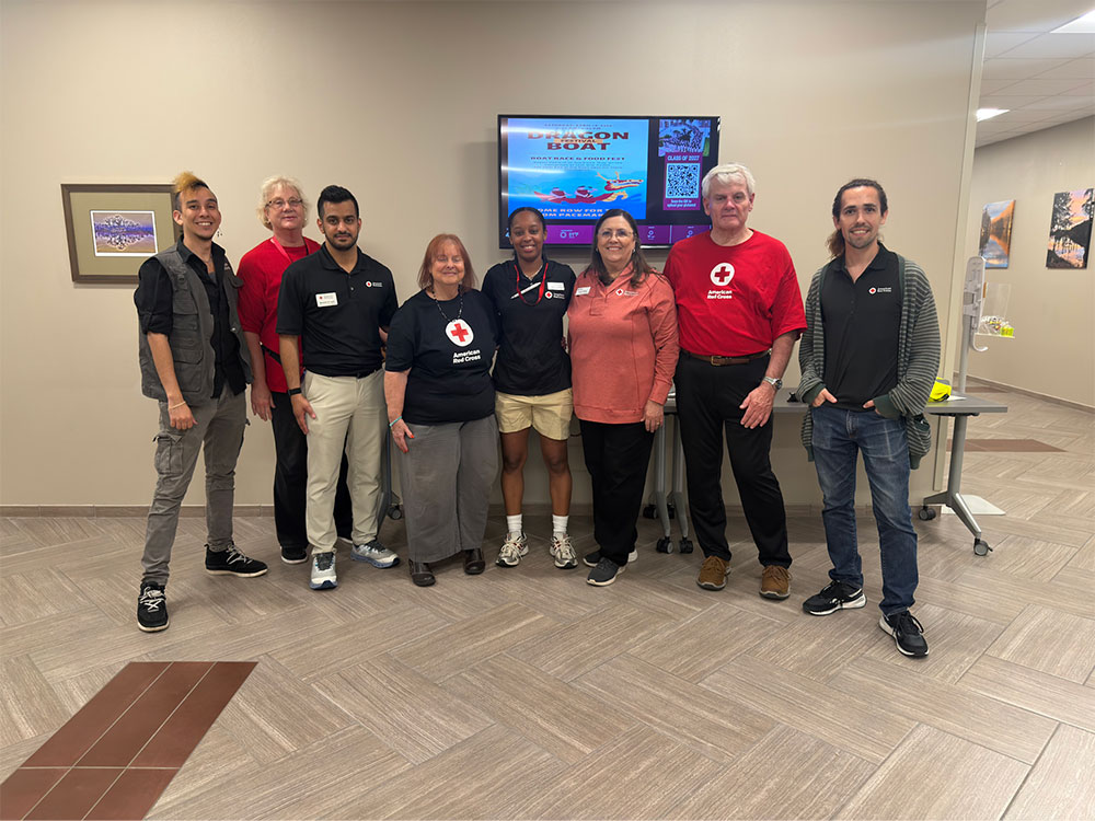 A group of eight people stands smiling in a room with a TV displaying a "Dragon Boat" graphic. They wear casual and Red Cross-themed attire.