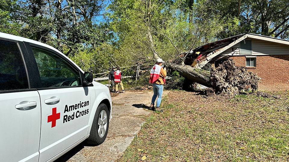 Red Cross volunteers and a Red Cross van next to a house with a fallen tree on part of the roof.