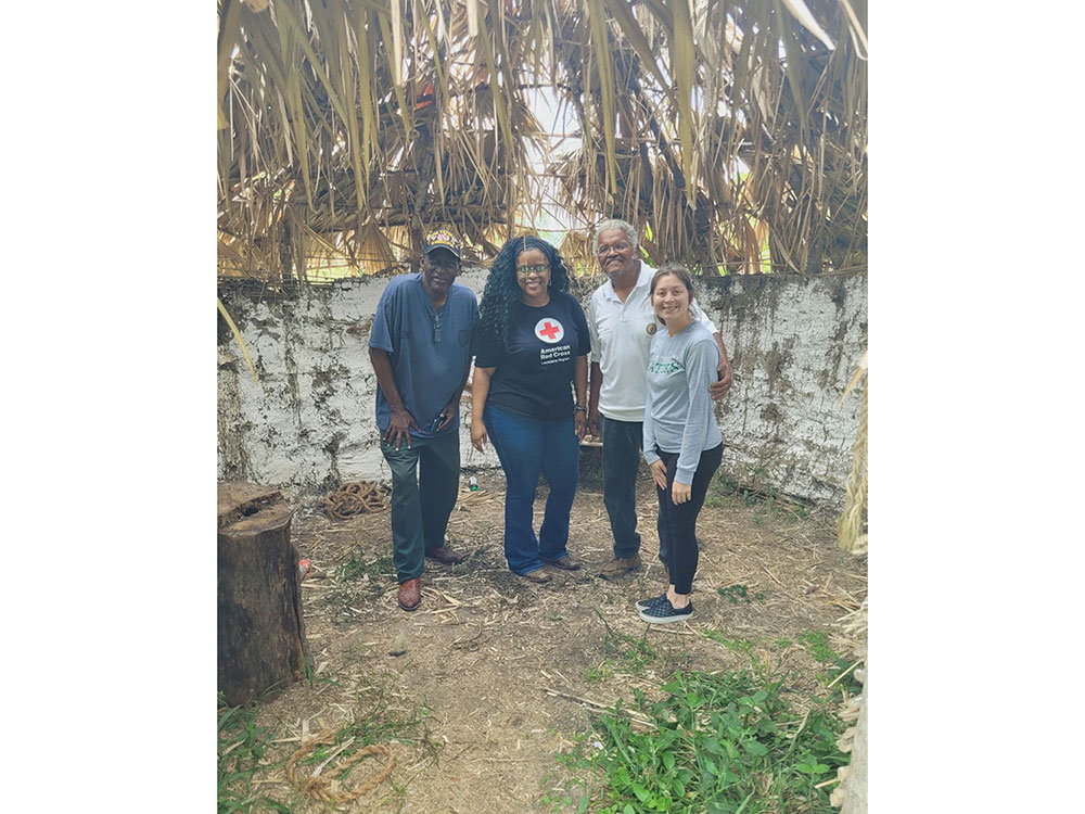 A group of four people stands in front of a hut, with a tree providing shade in the background.
