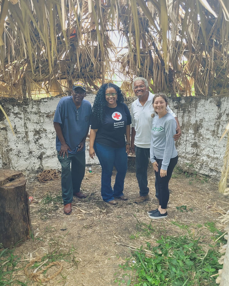 A group of four people stands in front of a hut, with a tree providing shade in the background.