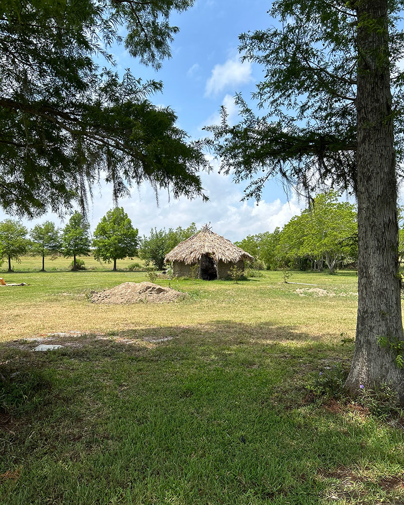 A small hut stands alone in the center of a vast green field under a clear blue sky.