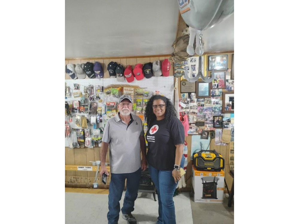 Two individuals stand in front of a wall filled with various tools, hats and pictures.