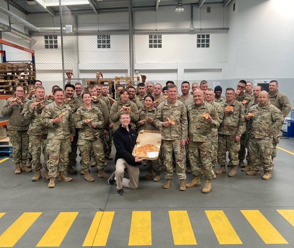 A group of soldiers holding slices of pizza and smiling, with a Red Cross volunteer.
