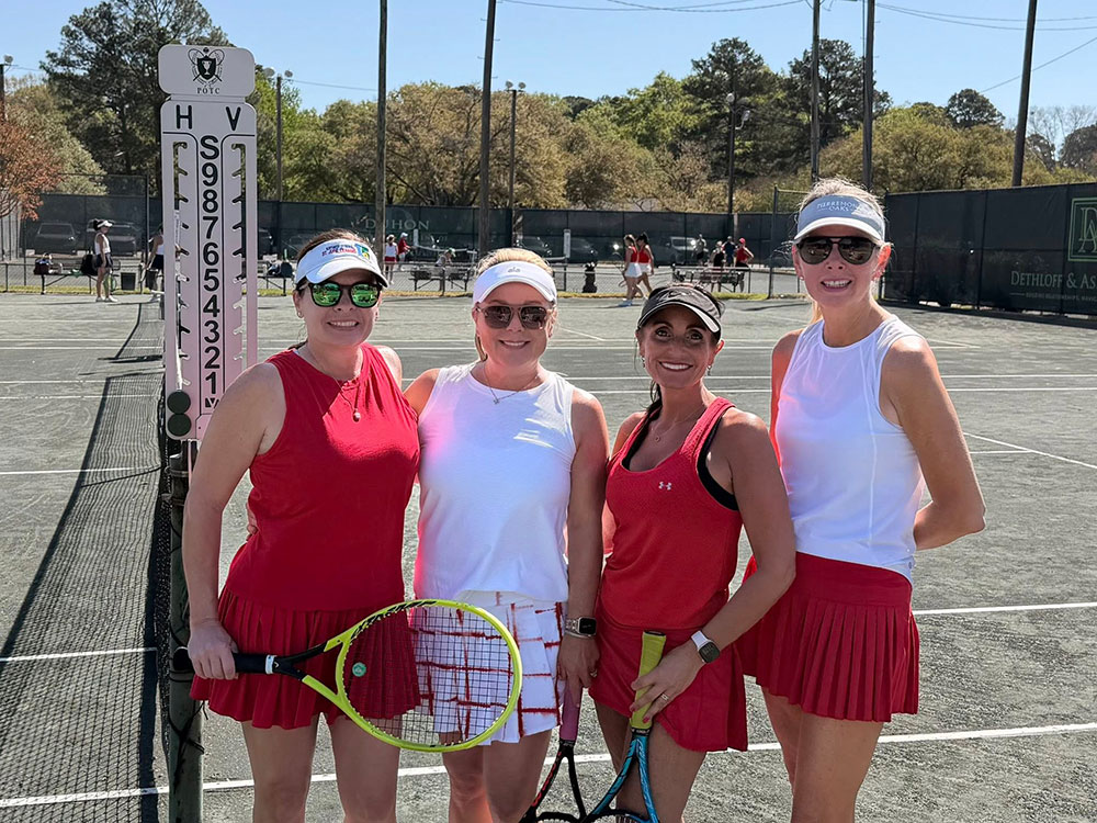 Four women in tennis attire pose happily on a sunny clay court, holding racquets. A score board is visible, with trees and a clear sky in the background.