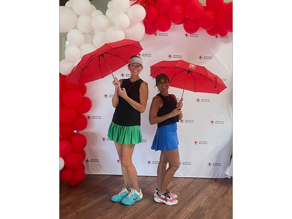 Two women, smiling and holding red umbrellas, pose in tennis outfits. They stand before a backdrop with the American Red Cross logo, framed by red and white balloons.