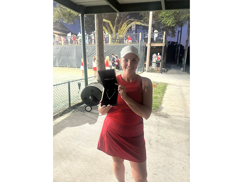 A woman in a red sports outfit and white cap holds a black box with a necklace, smiling. Tennis court background, evening event with festive lights.