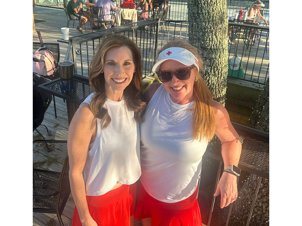 Two women smiling on a sunny deck, both wearing white tops and red skirts. One woman wears a Red Cross visor and sunglasses.
