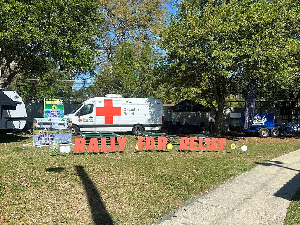 A disaster relief van with a large red cross is parked on grass alongside an RV. Red letters on the ground spell “Rally for Relief.” Trees provide shade.