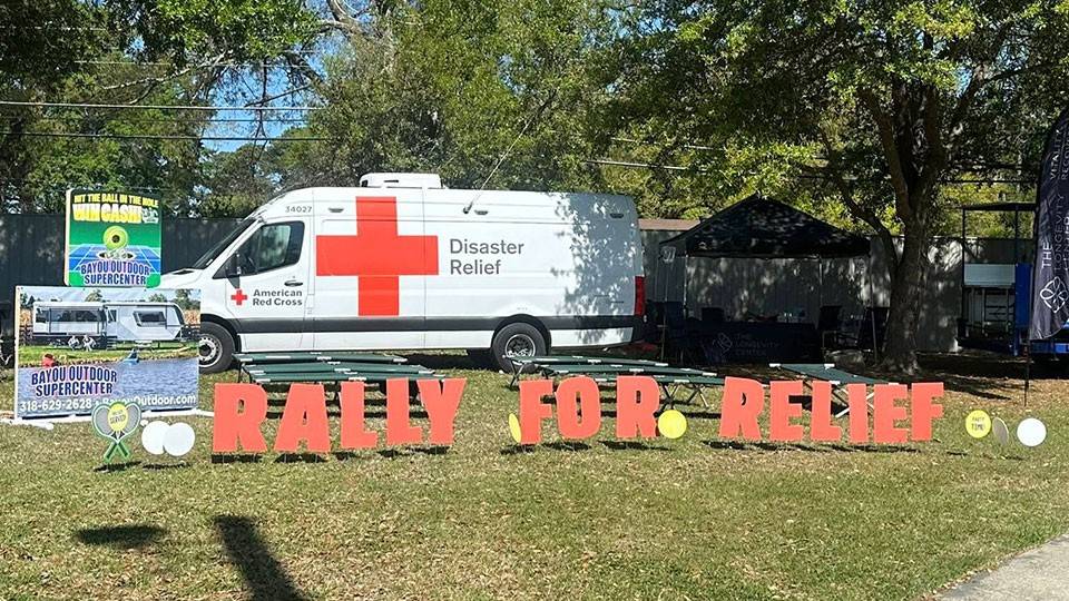 A disaster relief van with a large red cross is parked on grass alongside an RV. Red letters on the ground spell “Rally for Relief.” Trees provide shade.