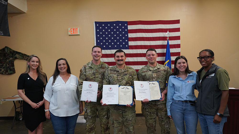 A group of three uniformed military personnel and four civilians stand smiling in front of a U.S. flag. The soldiers hold certificates.