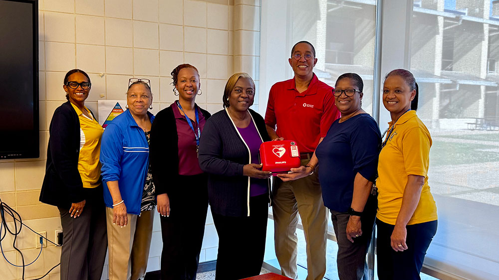 A group of seven smiling people stand indoors. The central woman holds a defibrillator box.