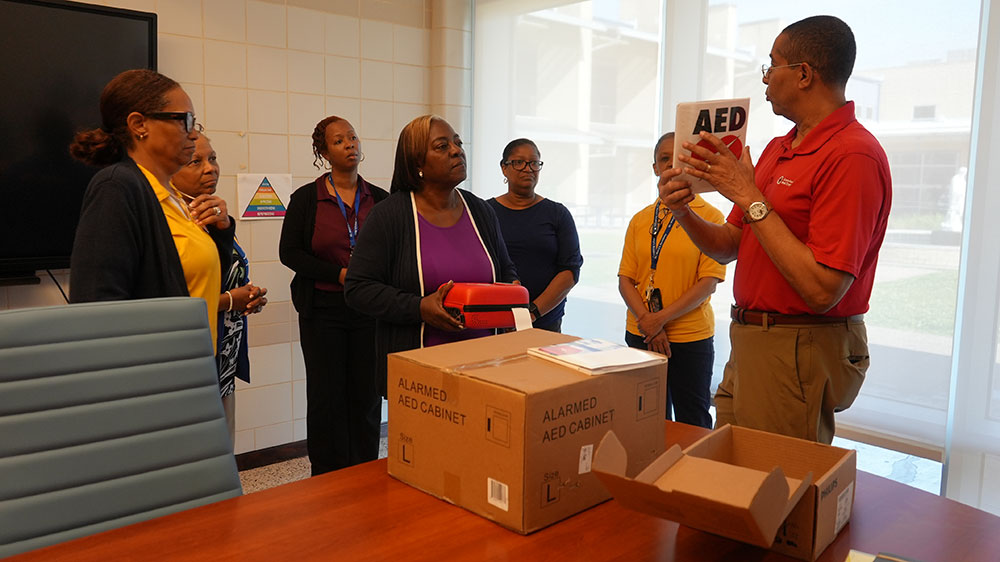A man in a Red Cross shirt conducts AED training for five attentive women in a conference room. He holds up an AED sign, as boxes are on the table.