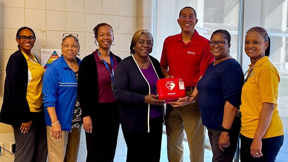 A group of seven smiling people stand indoors. The central woman holds a defibrillator box.