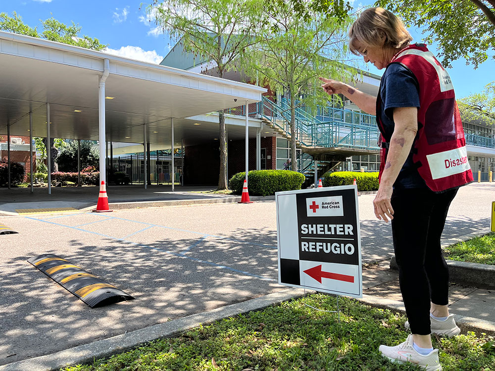 Red Cross volunteer standing next to a Red Cross shelter sign that's near a large building entrance.