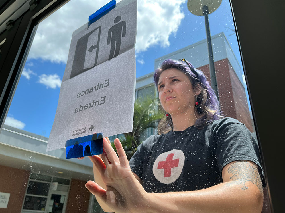 Red Cross volunteer taping a "Entrance/Entrada" sign on a window, wearing a Red Cross shirt. Outside view includes brick building and blue sky.