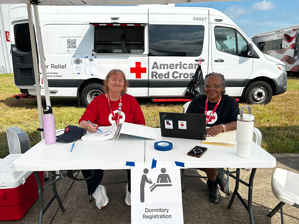 two Red Cross volunteers sitting at a table with paperwork and a laptop under a canopy.