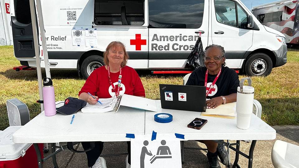 two Red Cross volunteers sitting at a table with paperwork and a laptop under a canopy.