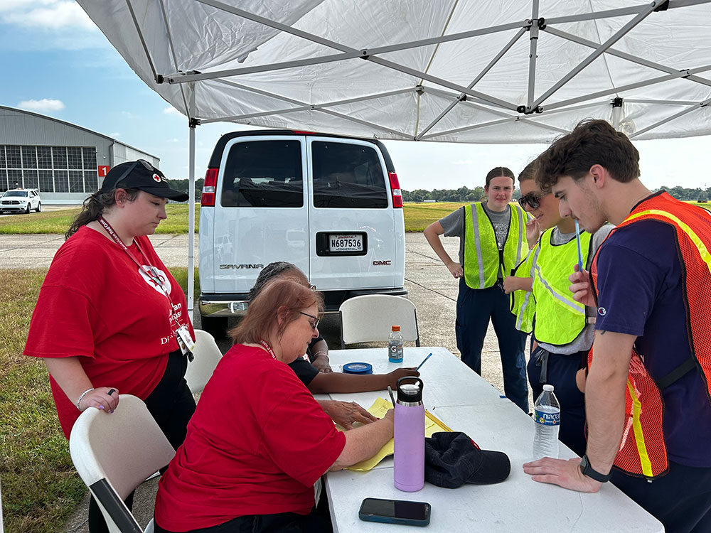 two Red Cross volunteers sitting and one standing at a table talking to three people that are wearing safety vests.