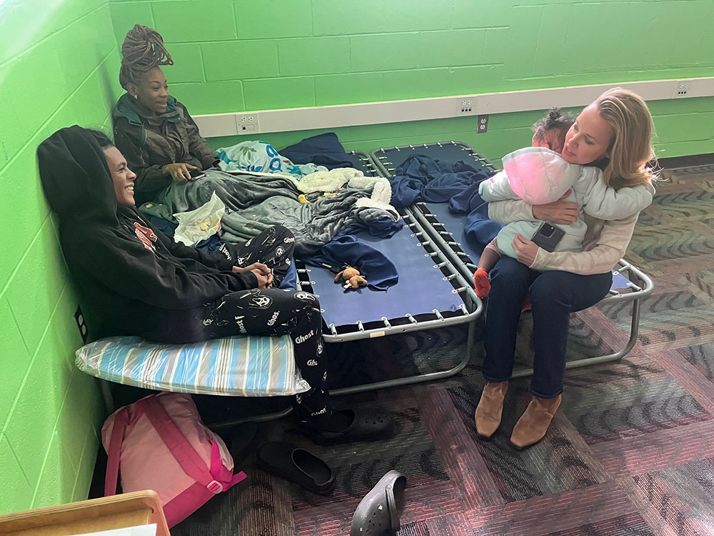 residents sitting on a cot in a shelter with a volunteer sitting next to them while holding their child.