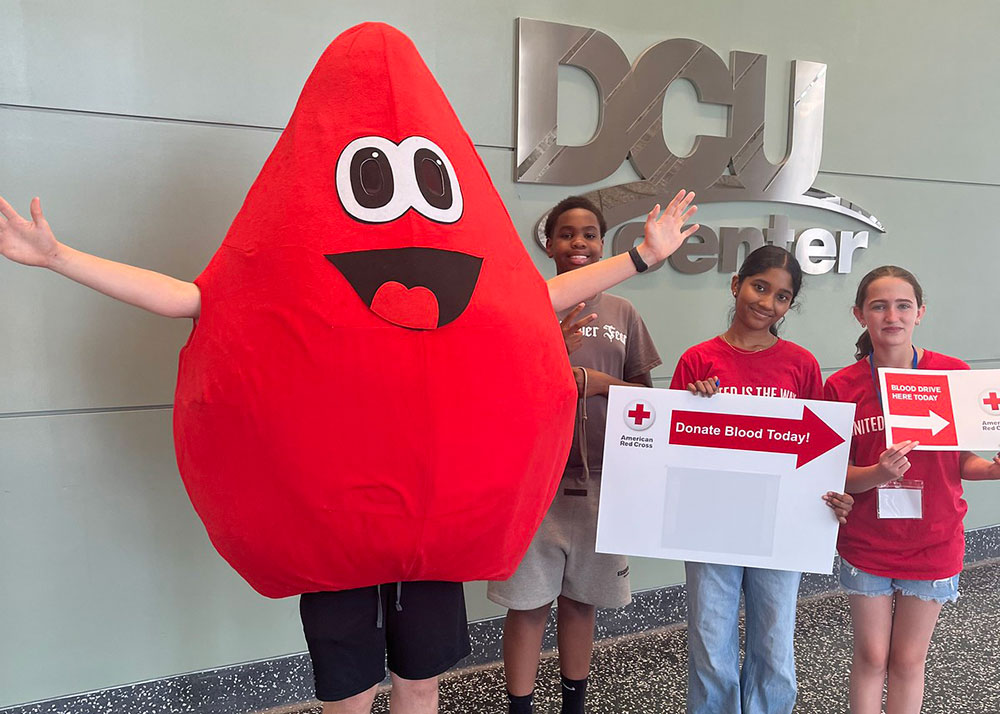Buddy the Blood Drop mascot next to three kids holding donate blood signs.