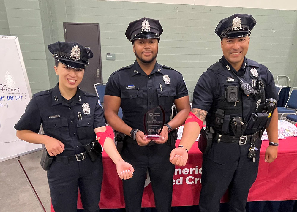 three police officers in uniform. One holding a battle of the badges award and the other two showing a red bandage around their arm.