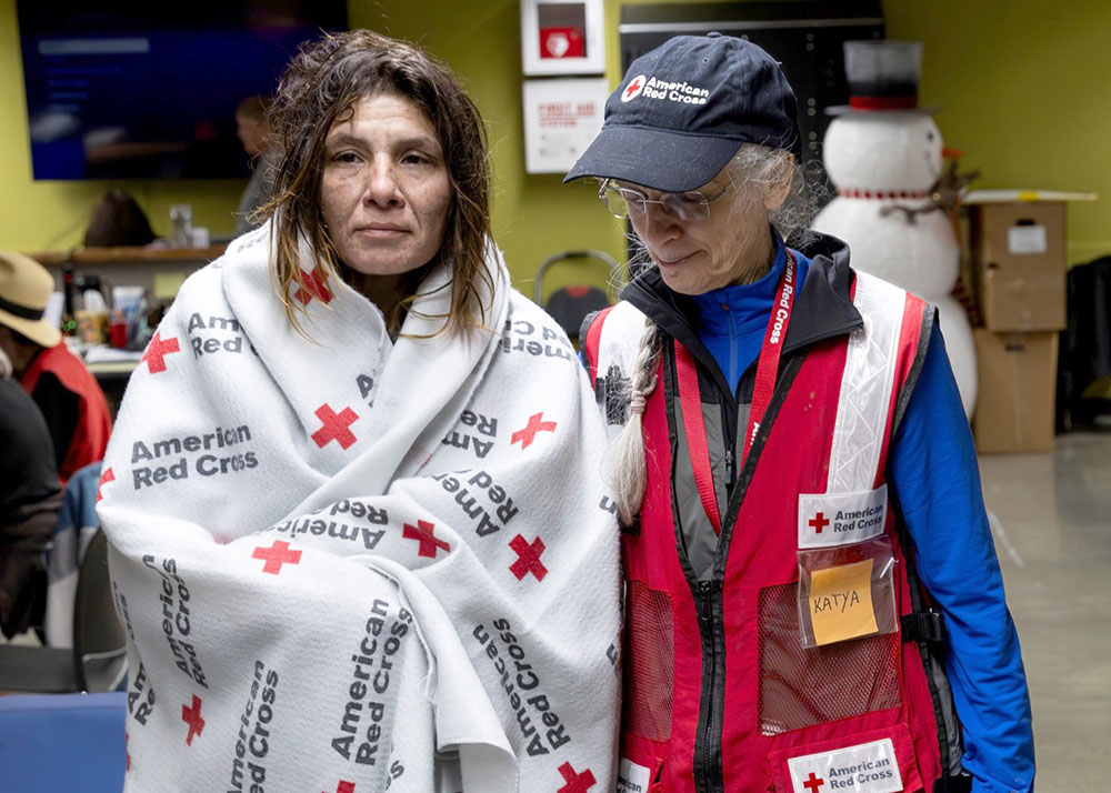 a Red Cross volunteer helping a person in a shelter that has a Red Cross blanket wrapped around their shoulders.
