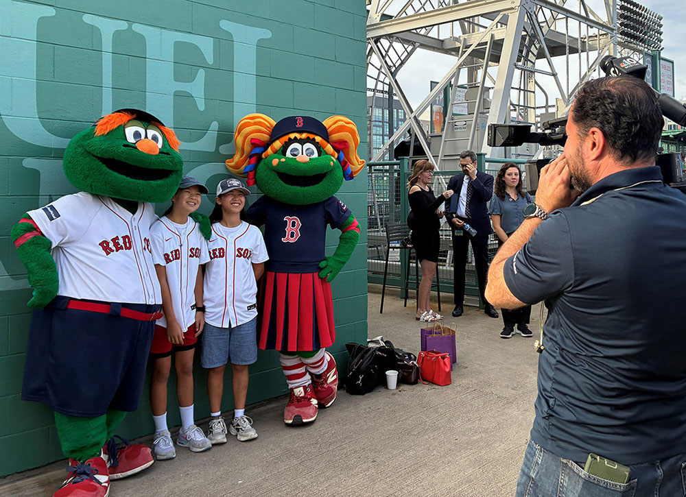 a woman and two kids with Red Sox shirts standing next to two Red Sox mascots with a guy holding a news camera.