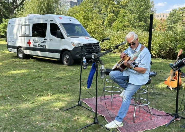 man sitting on stool playing guitar on a grassy field with a Red Cross van parked near by on the grass.